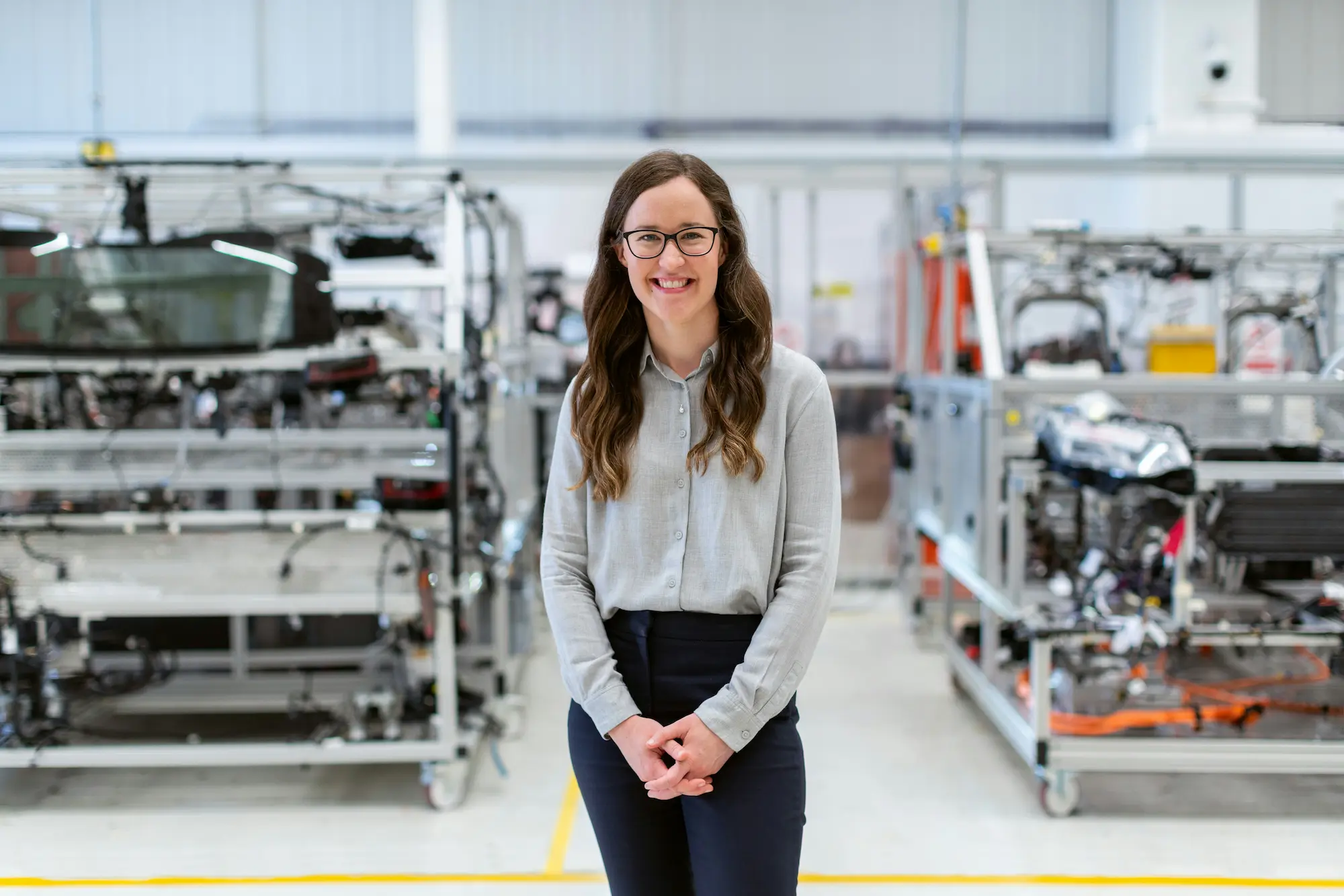 Woman standing facing the camera smiling. Some sort of manufacturing process behind her.